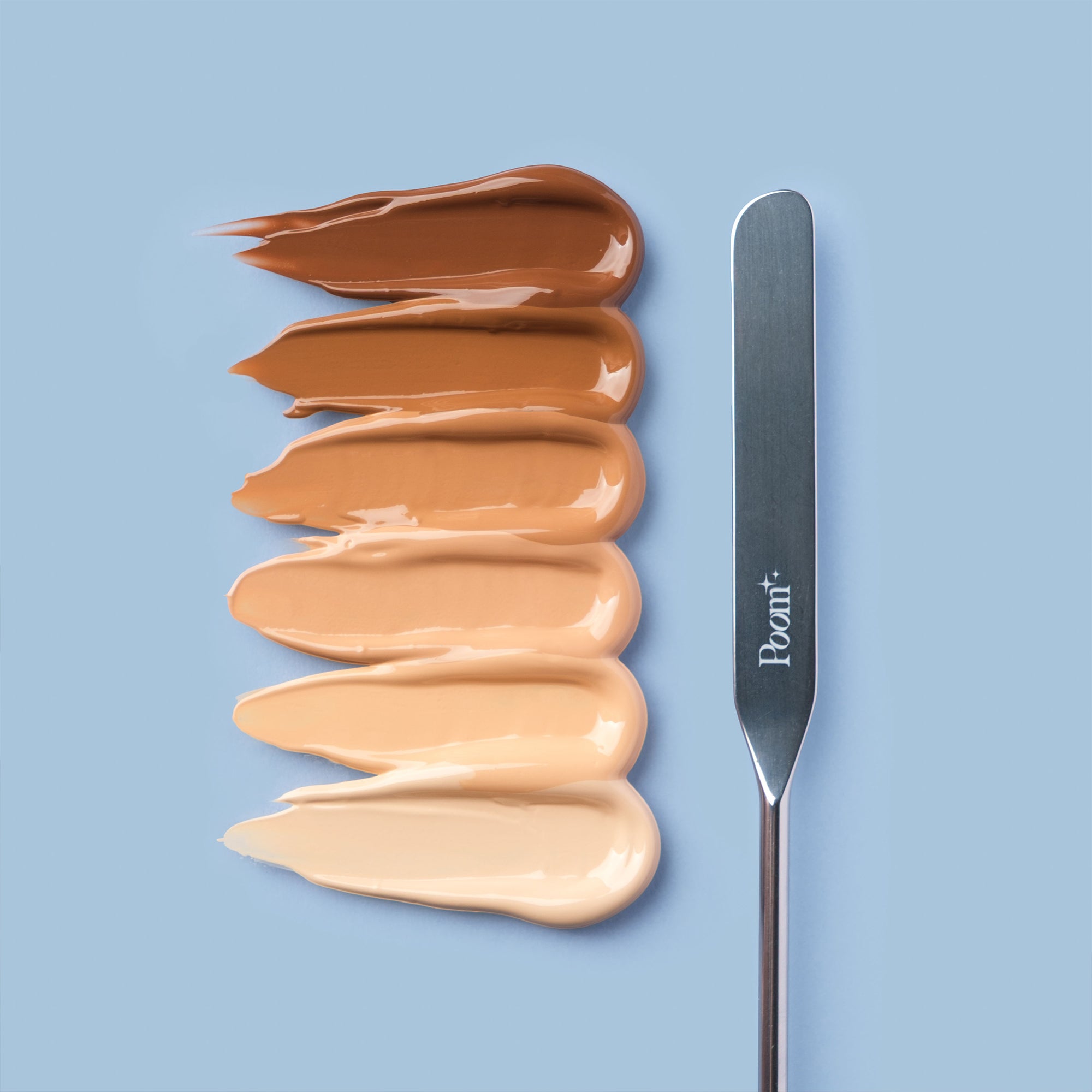 Row of swatches of 6 foundation shades with a spatula on a light blue background