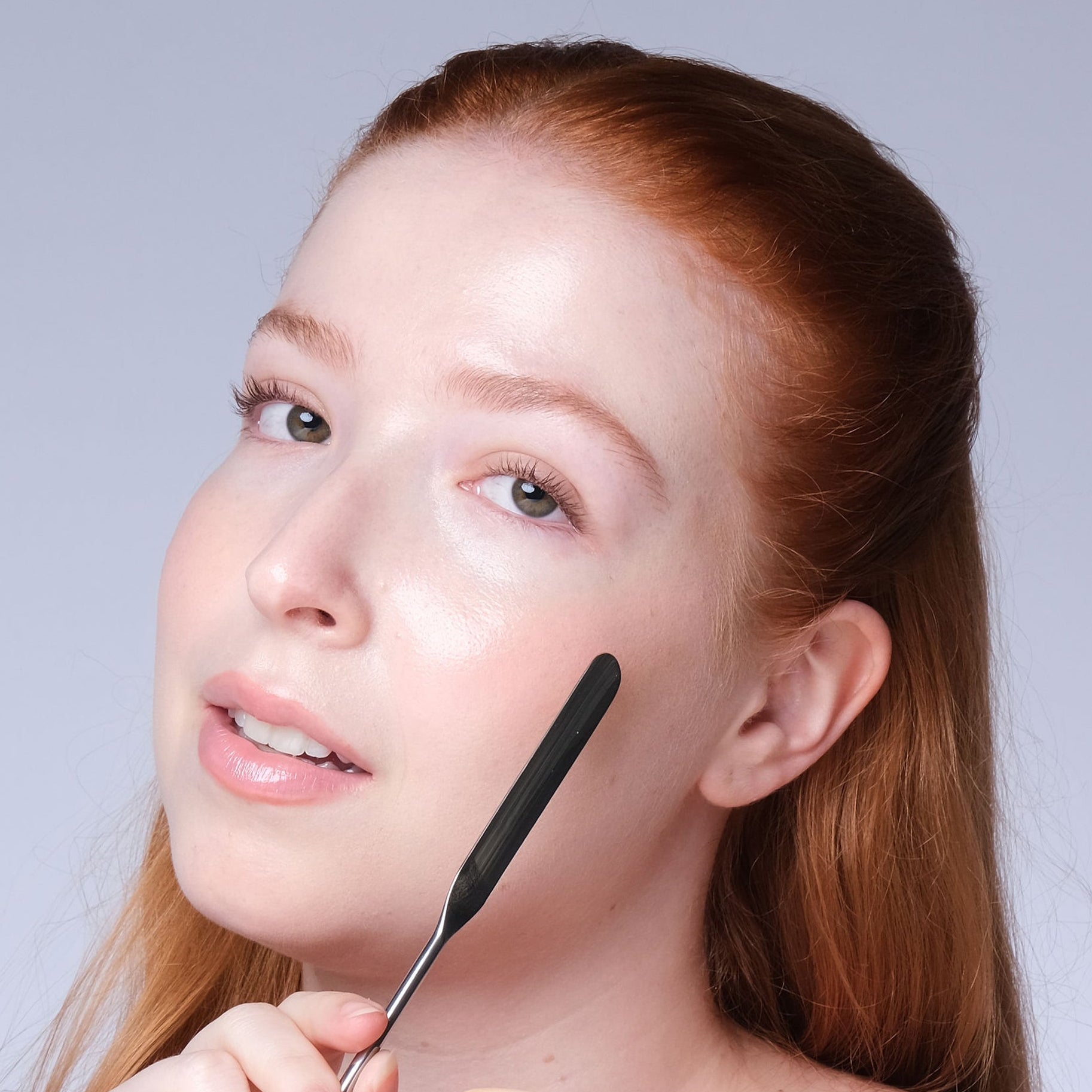 A woman with red hair applies a product using a black spatula and holds a white puff with the brand name 'Peach' visible, against a light gray studio background.